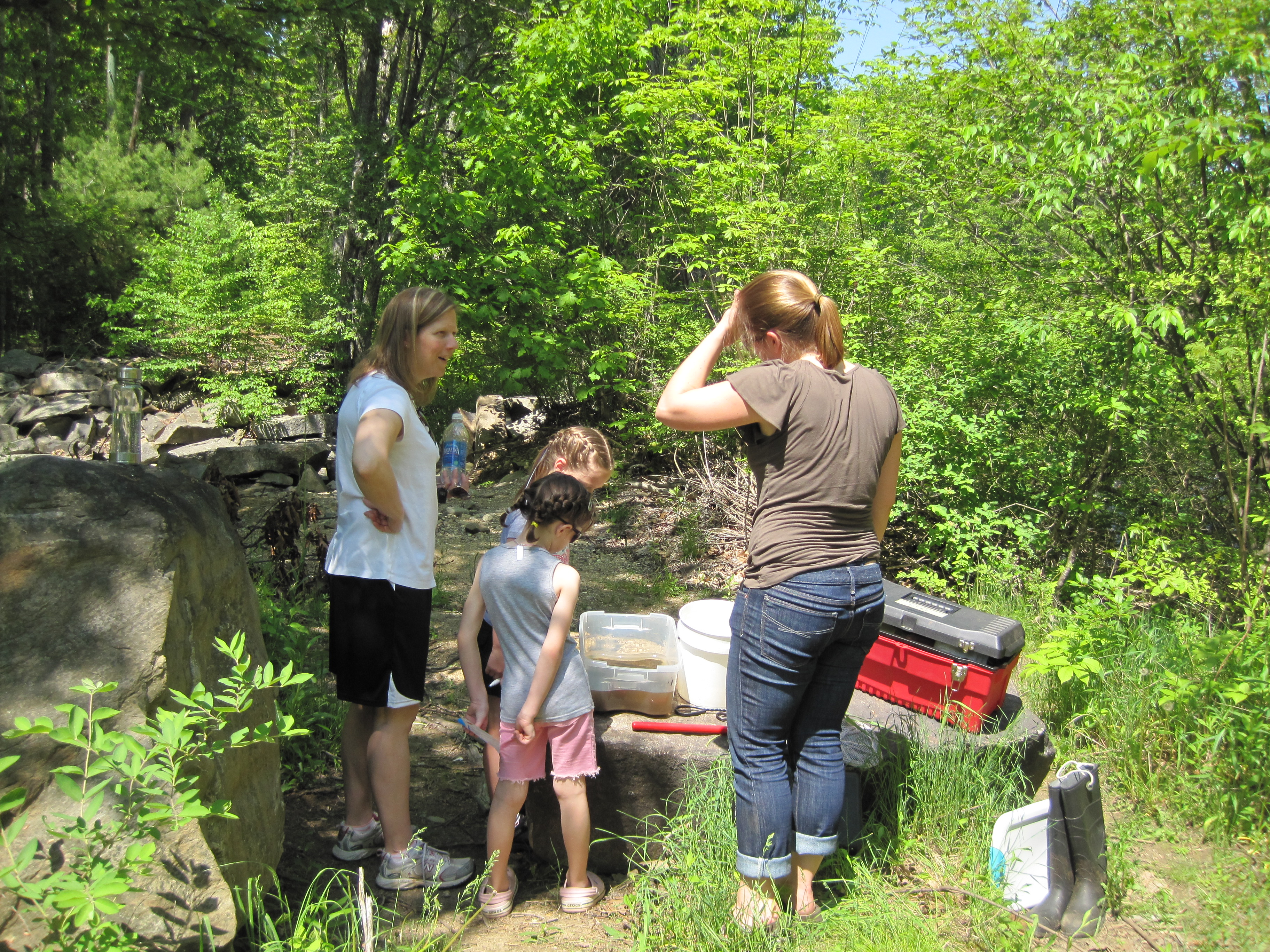 A group of 2 women and 2 children are standing along a bank on the Lamprey River. The group has an assortment of buckets and containers sitting on rocks.